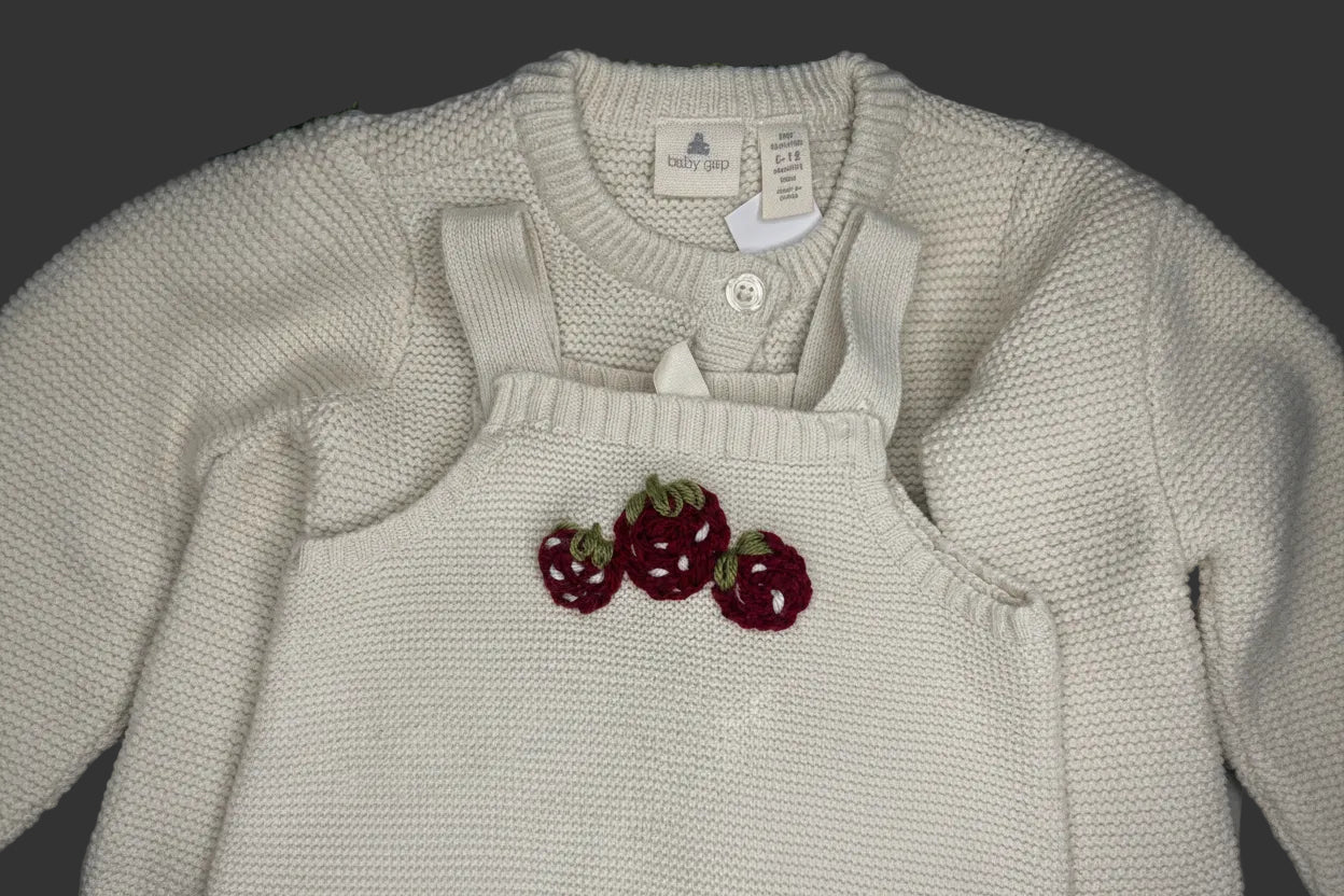 A close-up of a cream-colored knit baby sweater featuring three embroidered red strawberries with green tops on the chest, complete with a button closure and visible brand labels.